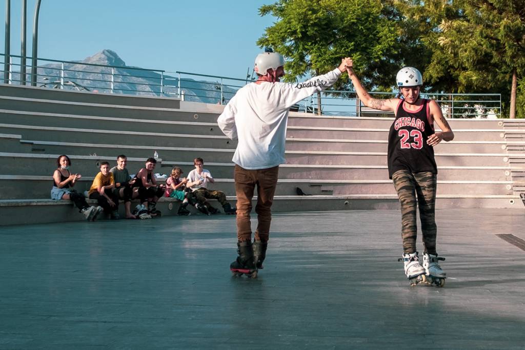 Two skaters holding hands on a plaza while the group watches from the steps